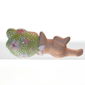 K&ouml;nnte beinhalten: Dekorative Figur einer liegenden Figur. Der Kopf ist mit einer hellgr&uuml;nen, juwelenbesetzten Bedeckung mit rosa und wei&szlig;en Blumenakzenten verziert. Der K&ouml;rper hat eine hellbraune Farbe.