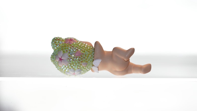 K&ouml;nnte beinhalten: Dekorative Figur einer liegenden Figur. Der Kopf ist mit einer hellgr&uuml;nen, juwelenbesetzten Bedeckung mit rosa und wei&szlig;en Blumenakzenten verziert. Der K&ouml;rper hat eine hellbraune Farbe.