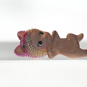 K&ouml;nnte beinhalten: Kleine, hellbraune Figur eines Babys, das liegt und einen mit rosa und goldenen Juwelen besetzten Hut tr&auml;gt. Die Figur hat gro&szlig;e Augen und liegt auf einer wei&szlig;en Oberfl&auml;che. Das Baby befindet sich in einer entspannten Pose.