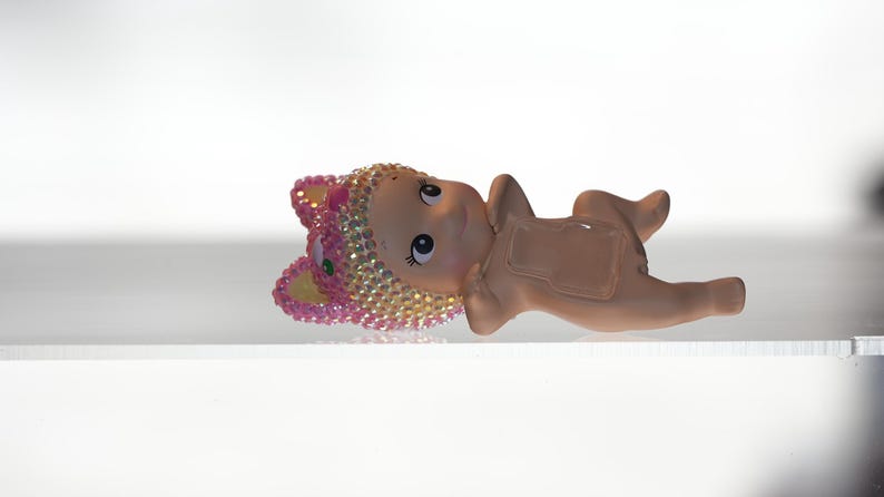 K&ouml;nnte beinhalten: Kleine, hellbraune Figur eines Babys, das liegt und einen mit rosa und goldenen Juwelen besetzten Hut tr&auml;gt. Die Figur hat gro&szlig;e Augen und liegt auf einer wei&szlig;en Oberfl&auml;che. Das Baby befindet sich in einer entspannten Pose.