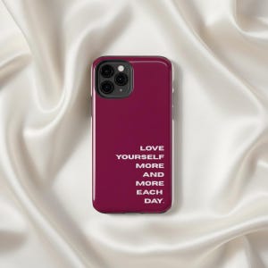 以下が含まれることがあります： 「LOVE YOURSELF MORE AND MORE EACH DAY」の白文字が入った、バーガンディ色の携帯電話ケース。携帯電話ケースには黒いカメラモジュールがあり、白いサテンの背景に置かれています。