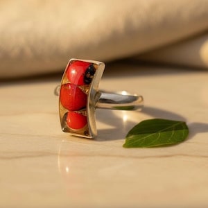May include: A silver ring with a rectangular setting containing three red, round beads. The setting has a gold-colored border. A green leaf rests nearby on a light-colored surface.