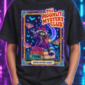 Puede incluir: Una camiseta negra con un gráfico de un mapache con sombrero y abrigo morados, sosteniendo una linterna y una pequeña pistola mientras está sentado sobre una pila de libros. El diseño incluye polillas, una luna creciente y el texto THE MOONLIT MYSTERY CLUB READ AFTER DARK.