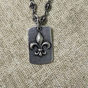 May include: A silver-toned metal necklace featuring a rectangular dog tag pendant with a raised fleur-de-lis emblem. The chain is composed of small, oval-shaped links, each decorated with a miniature fleur-de-lis motif, creating a cohesive design.