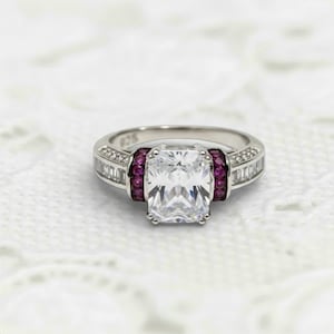 May include: A silver ring with a large, rectangular, clear gemstone at the centre. The ring features smaller clear stones and a row of small, round, red gemstones. The ring is set on a white, textured surface.