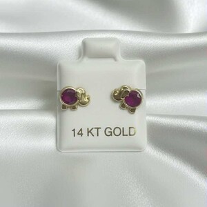 May include: A pair of gold elephant stud earrings with a round, magenta-colored stone in the center of each elephant. The earrings are displayed on a white card with the text "14 KT GOLD".