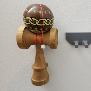 May include: A wooden kendama toy with a brown ball decorated with yellow chain and star patterns. The ball is connected to the wooden handle with an orange string. Two gray and brown wall-mounted shelves are also visible.