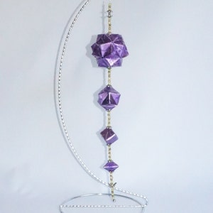 May include: A purple origami ornament hanging from a silver metal stand. The ornament is made of multiple geometric shapes and is suspended from a chain of gold beads.