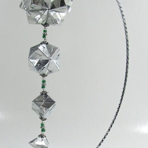 May include: A silver origami ornament with a geometric design hangs from a silver metal stand. The ornament is made of multiple folded pieces of paper and is decorated with green beads.