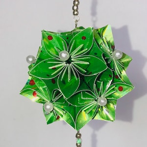 Pode incluir: Enfeite de origami verde feito à mão, com um design de estrela. O enfeite apresenta bolinhas vermelhas e pérolas brancas. Ele pende de uma corrente de contas prateada, perfeito para decorações festivas.