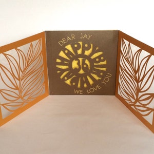 Puede incluir: Una tarjeta marrón con un diseño de hoja dorada en la parte delantera y trasera. El interior de la tarjeta tiene un círculo dorado con las palabras "Querido Jay Feliz Cumpleaños Te queremos" dentro.