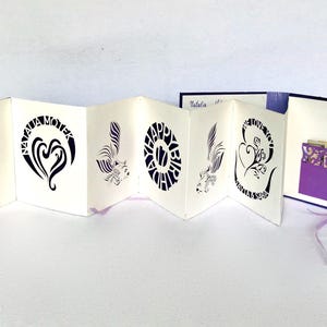 Puede incluir: Una tarjeta de felicitación blanca, estilo acordeón, con diseños gráficos y texto en negro. La tarjeta presenta un diseño de corazón, un mensaje de Feliz Cumpleaños y un bolsillo morado con recortes decorativos. Se adjunta una cinta morada.