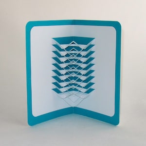 May include: A teal card with a white, geometric, 3D pop-up design. The design is made of overlapping, layered, white squares.