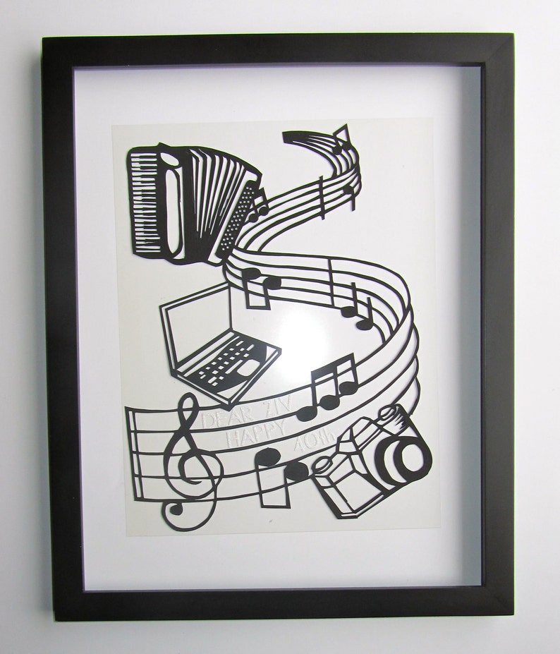 May include: Black and white paper cut art framed in a black frame. The art features a musical staff with notes, a camera, a laptop, and an accordion. The text "Dear Ziv Happy 40th" is visible on the staff.
