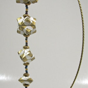 May include: A gold metal ornament stand with a white and gold origami sphere, a square, and a diamond shape hanging from it. The stand has a round base and a curved top. The ornament is decorated with beads.