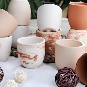 Könnte beinhalten: Eine Sammlung von Keramik-Blumentöpfen in verschiedenen Größen und Farben, darunter Weiß, Pfirsich und Terrakotta. Einige Töpfe haben ein Marmormuster. Dekorative geflochtene Kugeln in Braun und Weiß sind ebenfalls vorhanden.
