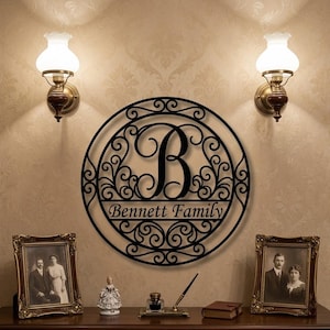 May include: Black metal family name sign with the letter "B" in the center, surrounded by decorative scrollwork. The words "Bennett Family" are displayed below the letter. Two wall sconces with white glass shades flank the sign.