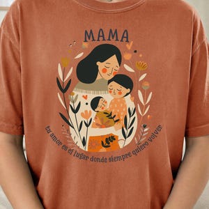 May include: Burnt orange t-shirt with the word "MAMA" above an illustration of a mother and two children embraced in a floral wreath. The text "Tu amor es el lugar donde siempre quiero volver" is below the illustration.