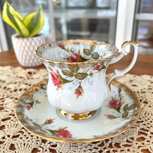 May include: A white teacup and saucer set with a floral rose pattern and gold trim. The teacup has a curved handle and sits on a matching saucer. The set is on a lace doily.