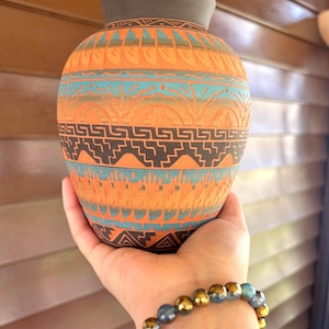 May include: A hand-painted ceramic vase with a dark gray rim and base. The body of the vase is orange with turquoise and black geometric patterns. The vase is held in a person's hand, with a beaded bracelet visible.