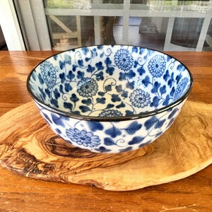 May include: A white and blue ceramic bowl with a floral pattern. The bowl has a black rim and is placed on a wooden cutting board. The floral design features blue flowers and leaves.