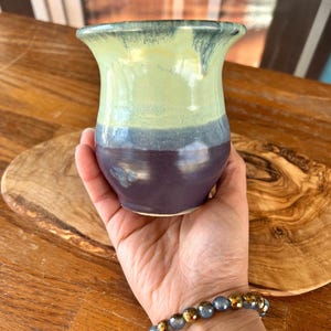 May include: A handmade ceramic vase with a flared rim. The vase features a color gradient, transitioning from light green at the top, through blue, to a deep purple at the base. The vase is held in a hand, showcasing its size and shape.