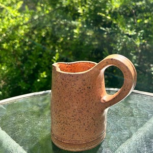 May include: A small, handmade ceramic pitcher with a rounded handle. The pitcher is a speckled brown color with a lighter interior. The pitcher is sitting on a glass surface with a blurred green background.
