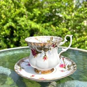 May include: A white teacup and saucer set with a floral rose design. The teacup has a gold rim and handle, and the saucer has a matching gold trim. The roses are pink and red with green leaves. The set is on a glass table.
