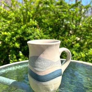 May include: A handmade ceramic mug with a cream-colored base and a curved handle. The mug features horizontal bands of gray, pink, and blue. The mug is sitting on a glass table with a green, leafy background.