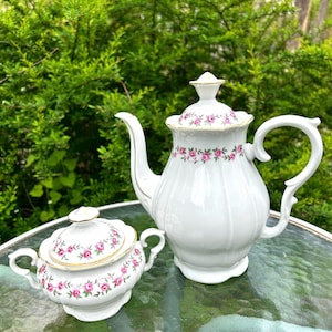 May include: A white porcelain teapot and a matching sugar bowl, both featuring a delicate pattern of small pink roses with green leaves and gold trim. The teapot has a tall, fluted body with a lid, while the sugar bowl has two side handles.