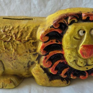 May include: A yellow ceramic money box shaped like a lion with a textured body, a red nose, and a mane featuring alternating black and orange segments. The lion has a horizontal coin slot on its back and a small red tail at the rear end.