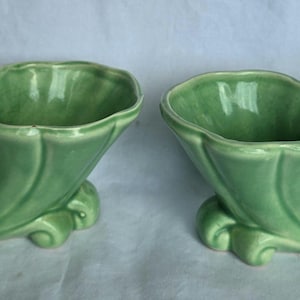 May include: A pair of matching light green ceramic planters, each featuring a sculpted squirrel figure attached to the side of a fluted, cornucopia-shaped vessel with a glossy finish and a curved base for stability on flat surfaces.