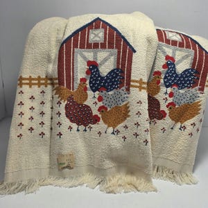 May include: Two off-white woven towels featuring a repeating pattern of a red barn, blue and brown chickens, and a brown fence. The towels have a fringed bottom edge and a small rectangular tag near the bottom corner with visible text.
