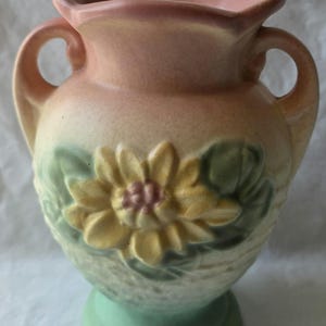 May include: A ceramic vase featuring a pastel pink rim and two side handles. The body displays a raised, embossed yellow sunflower with a pink centre, surrounded by green leaves against a textured, light green and cream-coloured base.