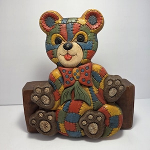 May include: A decorative bear figurine featuring a patchwork design with red, yellow, blue, and green segments. The bear has a red bow tie with blue polka dots, brown textured paws, and a light-coloured face with black eyes and a small red tongue.