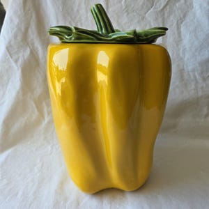 May include: A yellow ceramic canister shaped like a bell pepper with a glossy finish. The lid is designed as a green stem and leaves, featuring a smooth, reflective surface that mimics the natural contours and ridges of a fresh vegetable.