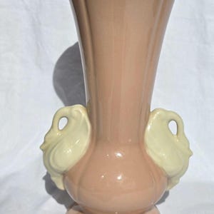 May include: A tall, glossy, light pink ceramic vase featuring a flared, scalloped rim and a rounded base. Two cream-colored swan-shaped handles are attached to the sides of the neck, providing a decorative accent to the smooth, solid-colored body.
