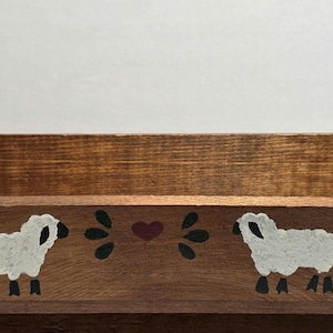 May include: A rectangular wooden wall shelf with a medium brown finish and rounded side panels. The front panel features a hand-painted design of two white sheep facing each other, separated by a small dark red heart and black leaf accents.