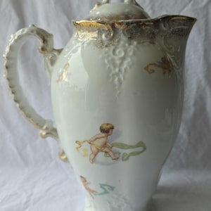 May include: A white porcelain pitcher with a lid, featuring gold-trimmed edges and a handle. The body is decorated with small, colorful illustrations of cherubs holding ribbons. The pitcher has a molded, textured surface pattern.