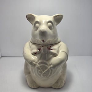 May include: A cream-colored ceramic cookie jar shaped like a seated animal with large ears. The figure wears a bow tie and holds a circular object. There are visible areas of red paint wear on the ears, nose, bow tie, and the circular object.