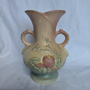 Puede incluir: Un jarrón de cerámica con acabado mate, cuerpo bulboso, borde acampanado y dos asas laterales. La superficie muestra un degradado en tonos beige y rosa, decorado con un relieve de una flor de color rosa y hojas de color verde.