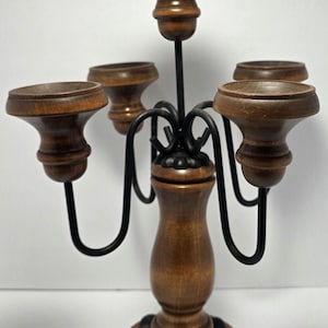 Puede incluir: Un candelabro de cinco brazos con un pilar central de madera y cuatro brazos curvos de metal negro. Cada brazo sostiene una copa de madera para velas con un acabado liso y pulido. La base es circular, hecha de madera con un detalle festoneado.