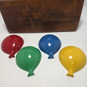 May include: Four glossy, balloon-shaped wall hooks in solid red, green, blue, and yellow. Each hook features a smooth, rounded top and a narrow base, designed to be mounted on a wall. The set is displayed in front of a dark brown wooden block.