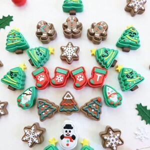 May include: A collection of Christmas-themed charms in various shapes, including Christmas trees, gingerbread men, snowflakes, stockings, and a snowman. The charms are in shades of green, brown, red, and white, with yellow star accents. The arrangement is on a white surface.
