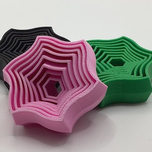 May include: Three nested star-shaped objects in black, pink, and green. Each object has a layered design with a central hexagonal opening. The objects are arranged on a white surface, showcasing their three-dimensional form.