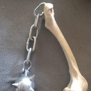May include: A medieval-style mace with a bone handle and a spiked metal ball. The handle is connected to the ball with a silver chain. The bone is off-white, and the metal ball is silver.
