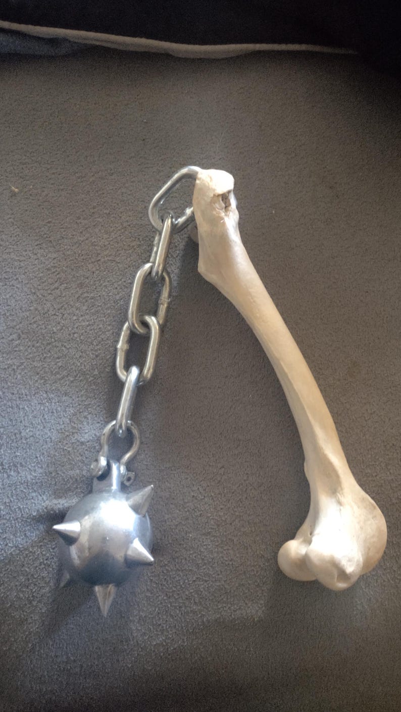 May include: A medieval-style mace with a bone handle and a spiked metal ball. The handle is connected to the ball with a silver chain. The bone is off-white, and the metal ball is silver.
