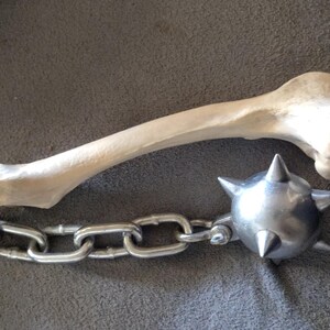 May include: A medieval-style mace with a bone handle and a spiked metal ball. The handle is a light beige color, and the ball is silver. The mace is connected to the handle with a silver chain.
