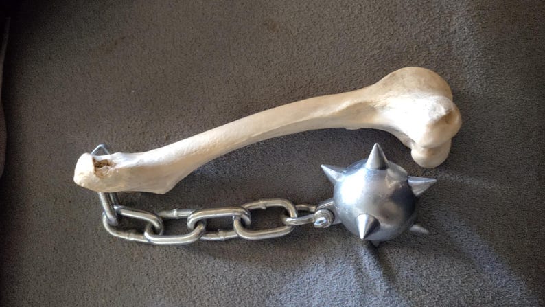 May include: A medieval-style mace with a bone handle and a spiked metal ball. The handle is a light beige color, and the ball is silver. The mace is connected to the handle with a silver chain.