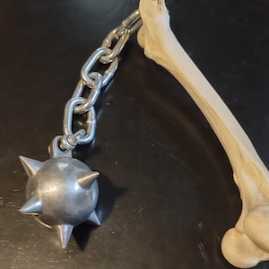 May include: A medieval-style mace with a spiked metal ball attached to a chain, next to a light-colored bone. The mace and bone are set against a dark background. The mace is a weapon.